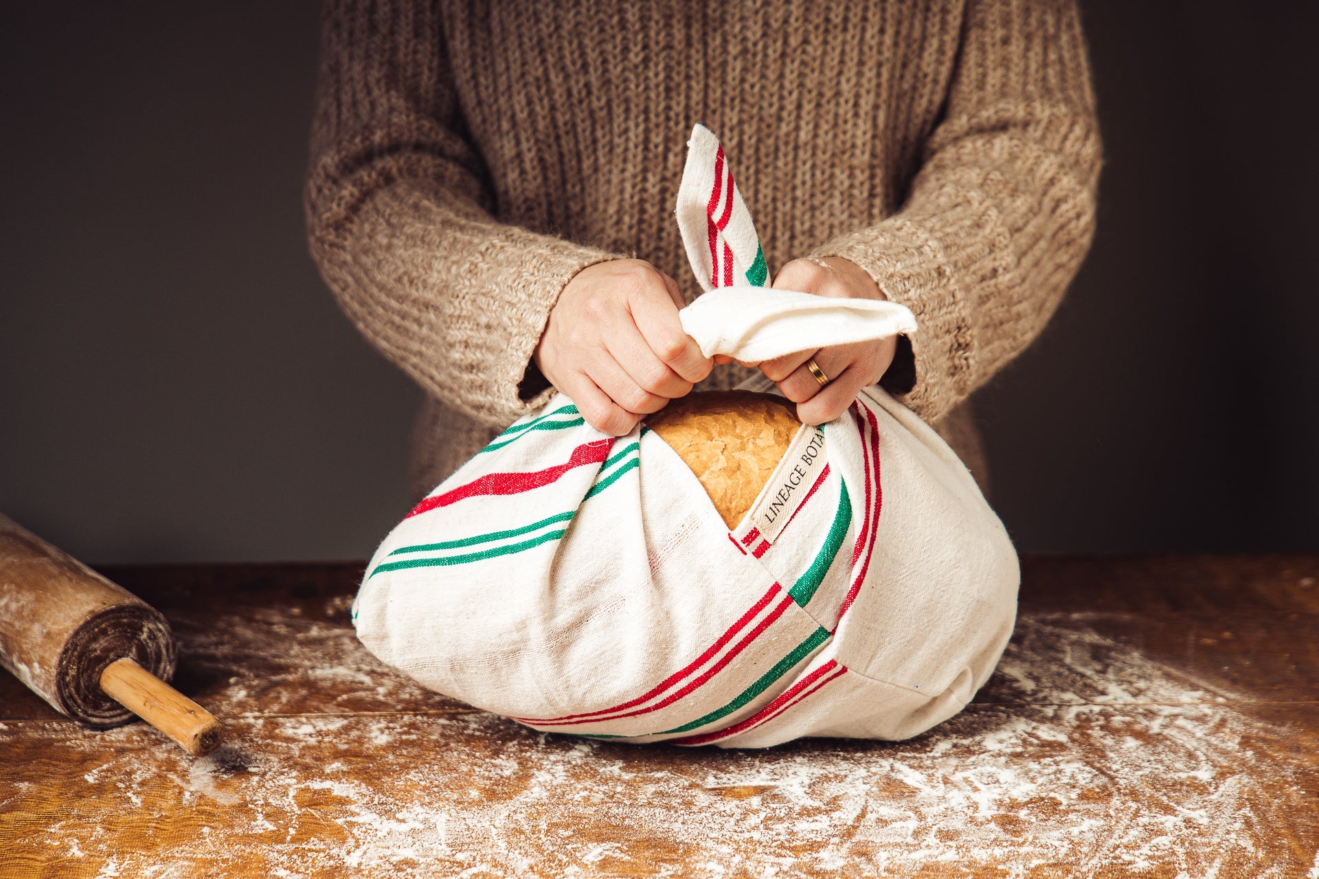 Antique handwoven Bulgarian cotton bag with green and red stripes tied around a loaf of bread on a flour-dusted surface.